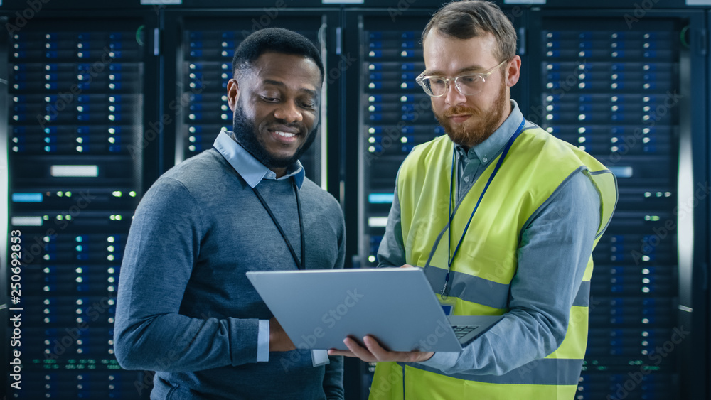 Poster Bearded IT Specialist in Glasses and High Visibility Vest with a Laptop Computer and Black Technician Colleague Talking in Data Center while Standing Next to Server Racks. Running Maintenance. 