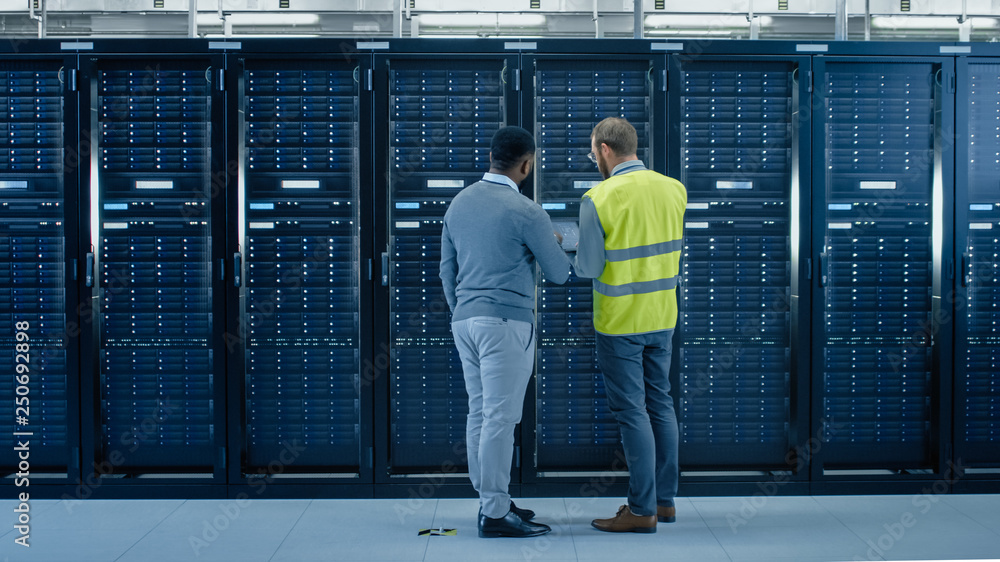 Aufkleber Bearded IT Specialist in Glasses and High Visibility Vest with a Laptop Computer and Black Technician Colleague Talking in Data Center while Standing Next to Server Racks. Running Maintenance. 