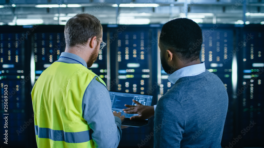 Poster Back View on Bearded IT Engineer in Glasses and High Visibility Vest with a Laptop Computer and Black Technician Colleague Talking in Data Center while Standing Next to Server Racks.