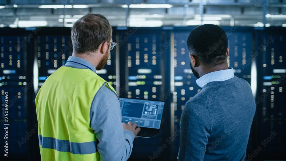 Poster Back View on Bearded IT Engineer in Glasses and High Visibility Vest with Laptop Computer and Black Technician Colleague Talking in Data Center while Standing Next to Server Racks. Running Maintenance