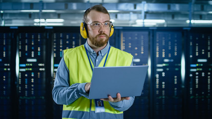 Bearded IT Specialist in Glasses and Headphones, wearing High Visibility Vest is Working on Laptop in Data Center Next to Server Racks.