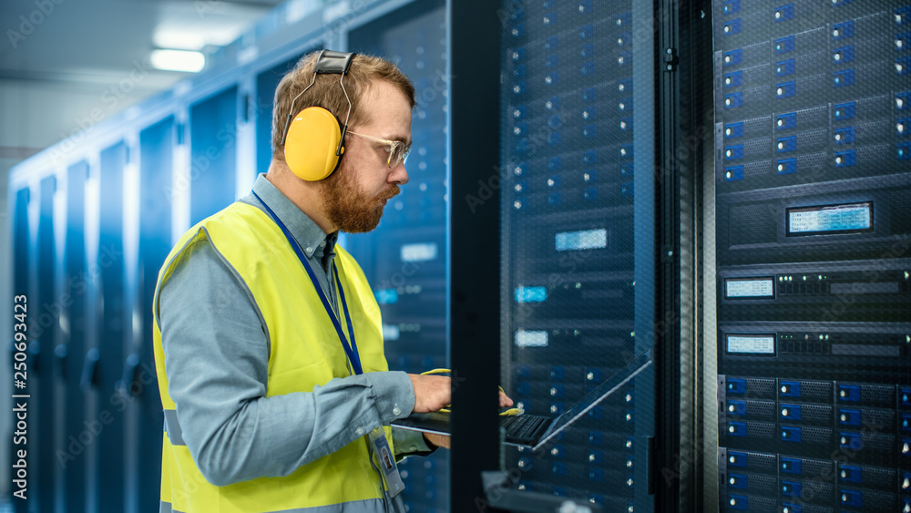 Poster IT Engineer in High Visibility Vest is Working on Laptop in Data Center while Standing Before Server Rack. Running Diagnostics or Doing Maintenance Work.