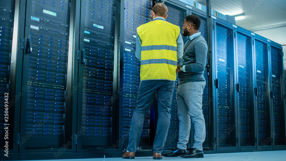 Poster IT Administrator with a Laptop Computer in High Visibility Vest and Black Male Engineer are Talking in Data Center while Standing Before a Server Rack. Running Diagnostics or Doing Maintenance Work. 
