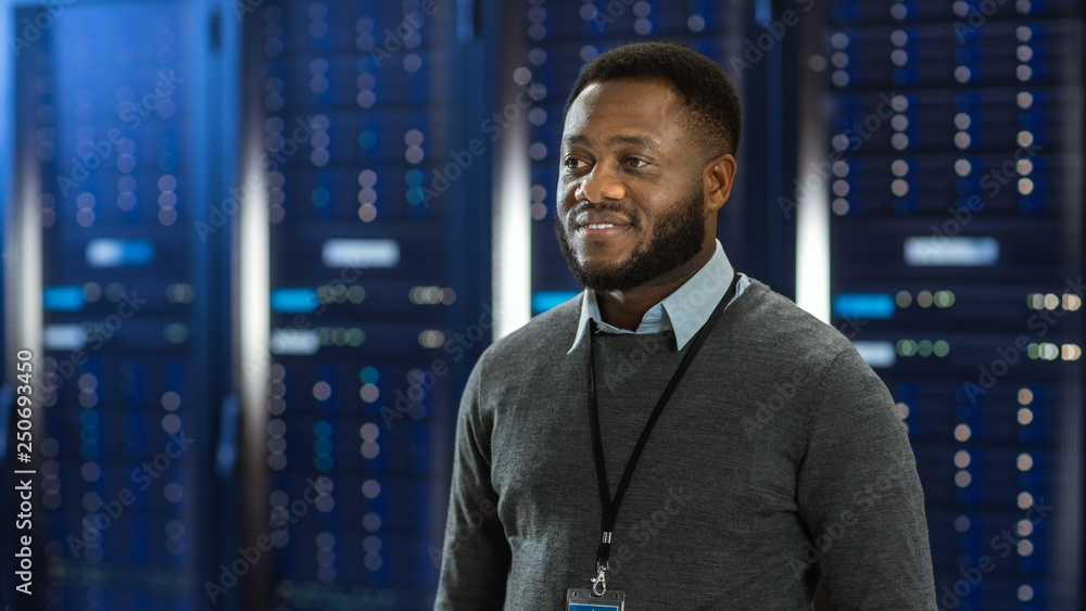 Poster Black Data Center IT Technician Standing in the Middle of a Server Rack Corridor. He is Smiling.