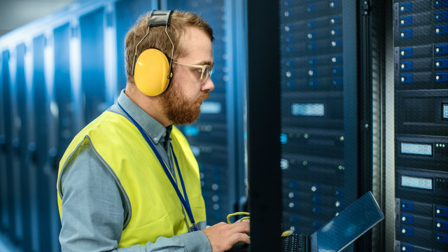 Bearded IT Specialist in Glasses and Headphones, wearing High Visibility Vest is Working on Laptop in Data Center Next to Server Racks. Running Diagnostics or Doing Maintenance Work.