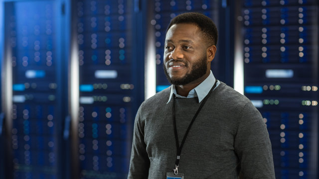 Black Data Center IT Technician Standing in the Middle of a Server Rack Corridor. He is Smiling.