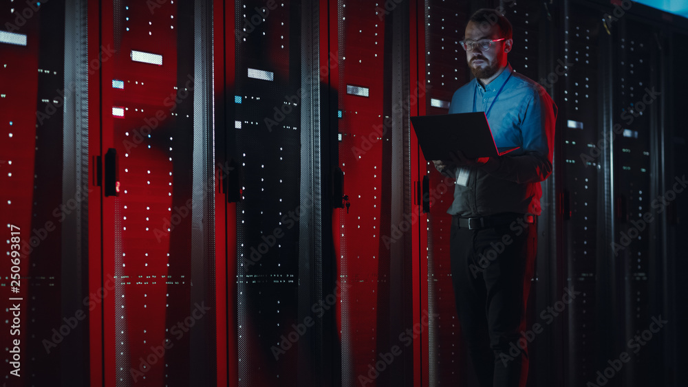 Leinwandbilder Bearded IT Specialist in Glasses is Working on Laptop in Data Center while Standing Near Server Rack. Running Diagnostics, Doing Maintenance Work. Emergency Red Light from Side Illuminating Specialist