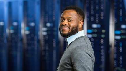 Young Black Data Center IT Technician Standing in Server Rack Corridor and Smiling to the Camera.