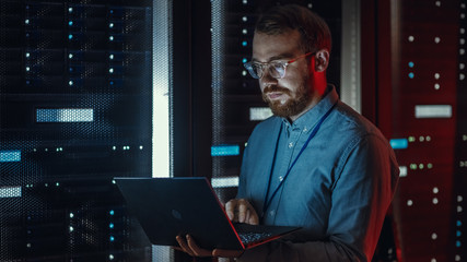 Bearded IT Specialist in Glasses is Working on Laptop in Data Center while Standing Near Server...