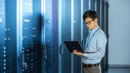 In the Modern Data Center: IT Engineer Standing Beside Server Rack Cabinets, Does Wireless Maintenance and Diagnostics Procedure with a Laptop. - obrazy, fototapety, plakaty
