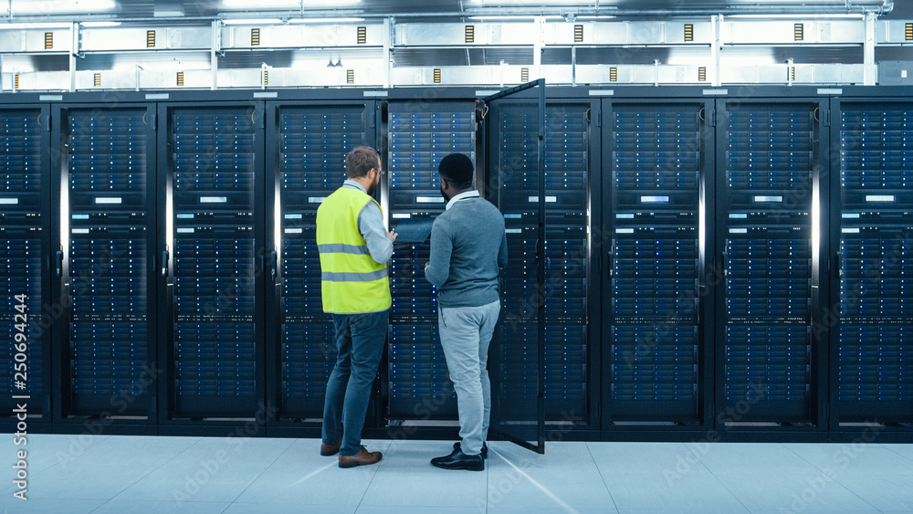 Fototapete IT Administrator with a Laptop Computer in High Visibility Vest and Black Male Engineer are Talking in Data Center while Standing Before a Server Rack. Running Diagnostics or Doing Maintenance Work. 