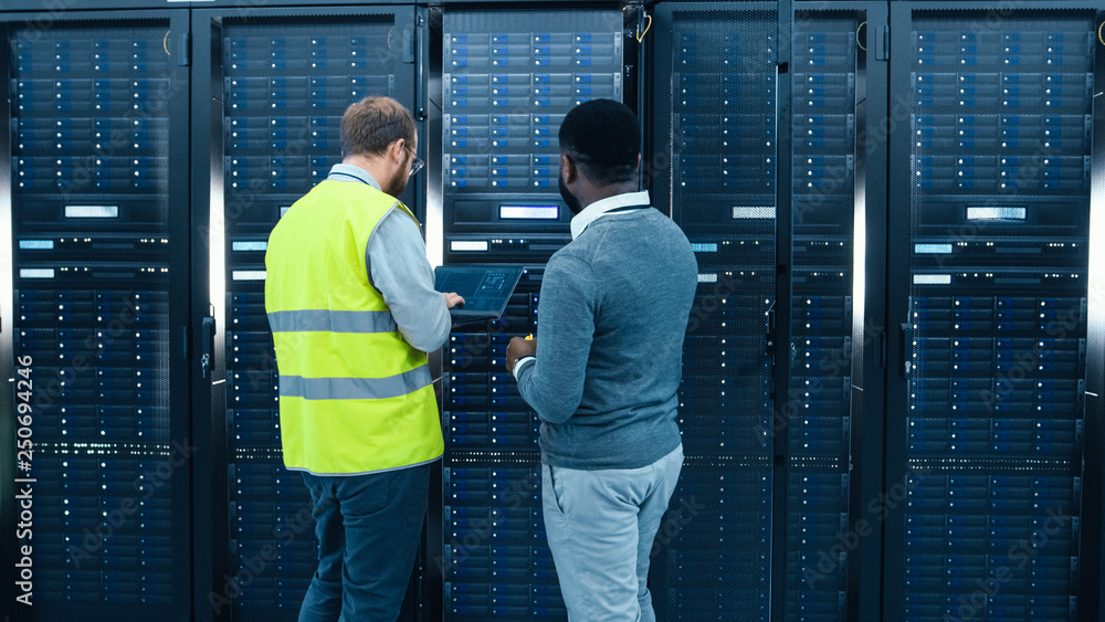 Aufkleber IT Administrator with a Laptop Computer in High Visibility Vest and Black Male Engineer are Talking in Data Center while Standing Before a Server Rack. Running Diagnostics or Doing Maintenance Work. 
