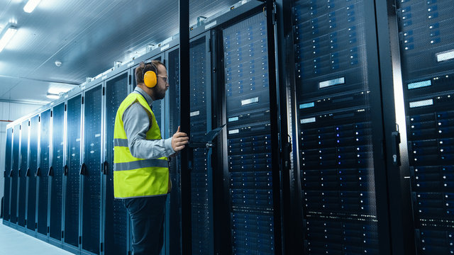 IT Administrator in High Visibility Vest is Opening the Door to the Server Rack. He's Working on Laptop in Data Center while Standing Before Them. Running Diagnostics or Doing Maintenance Work. 