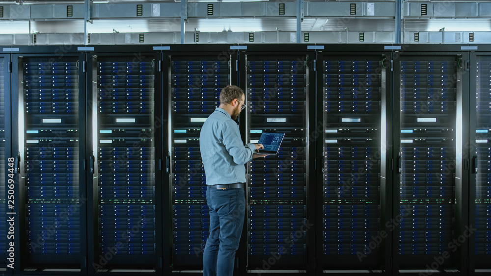 Aufkleber Bearded IT Specialist in Glasses is Working on Laptop in Data Center while Standing Before Server Rack. Running Diagnostics or Doing Maintenance Work.