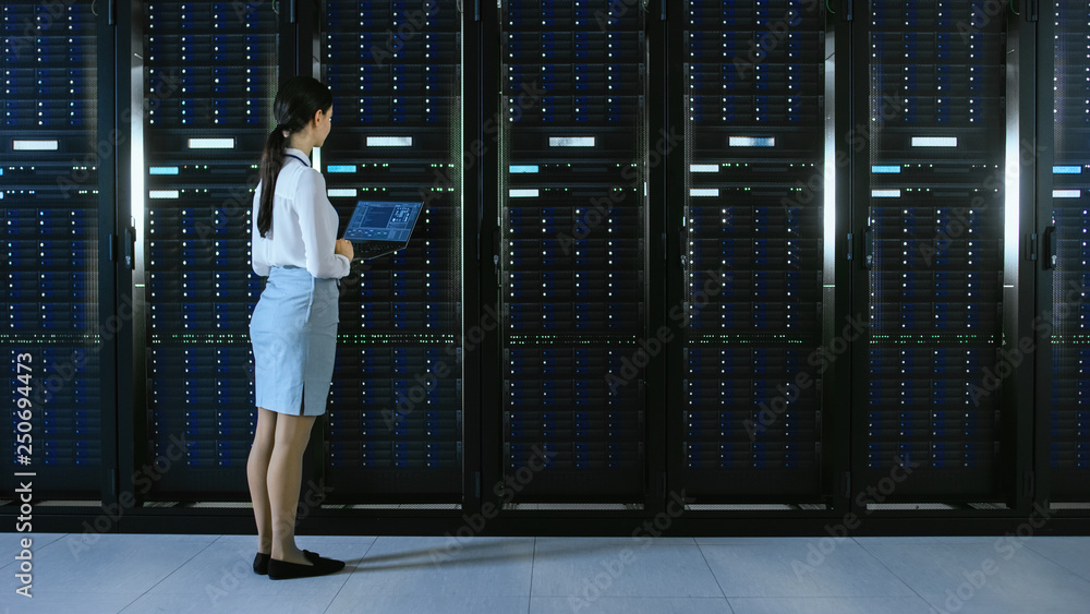 Poster Beautiful Data Center Female IT Technician Walking Through Server Rack Corridor with a Laptop Computer. She Stops and Visually Inspects Working Server Cabinets.