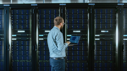 Bearded IT Specialist in Glasses is Working on Laptop in Data Center while Standing Before Server...