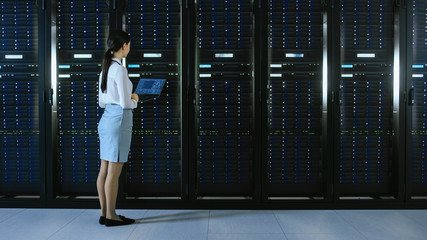 Beautiful Data Center Female IT Technician Walking Through Server Rack Corridor with a Laptop Computer. She Stops and Visually Inspects Working Server Cabinets.