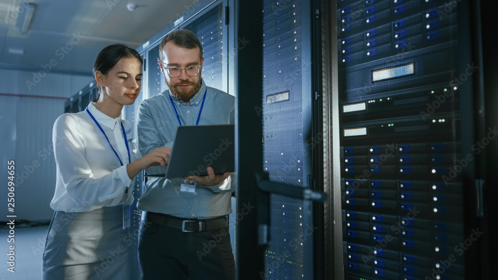 Aufkleber Bearded IT Technician in Glasses with a Laptop Computer and Beautiful Young Engineer Colleague are Talking in Data Center while Working Next to Server Racks. Running Diagnostics or Doing Maintenance
