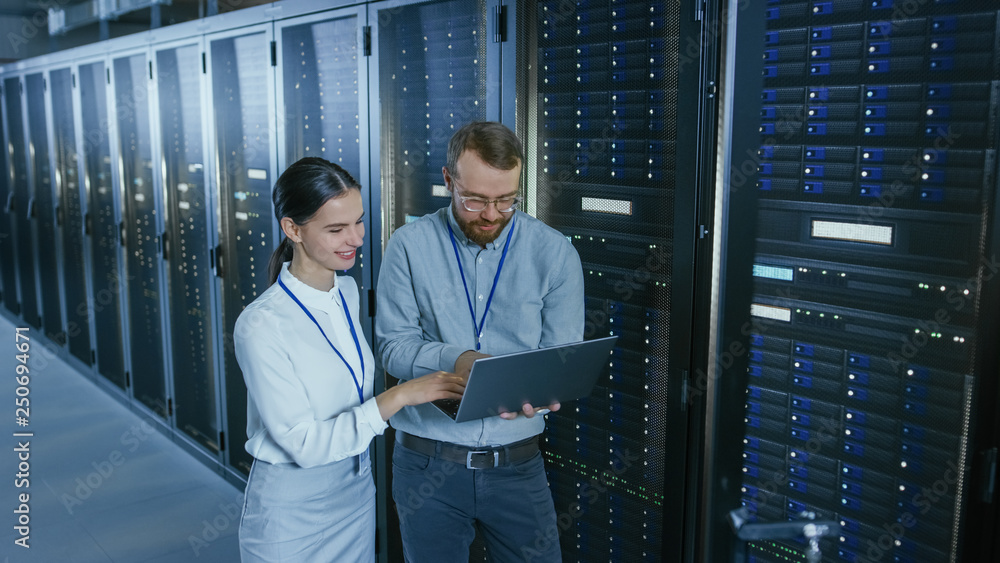 Poster Bearded IT Technician in Glasses with a Laptop Computer and Beautiful Young Engineer Colleague are Talking in Data Center while Working Next to Server Racks. Running Diagnostics or Doing Maintenance