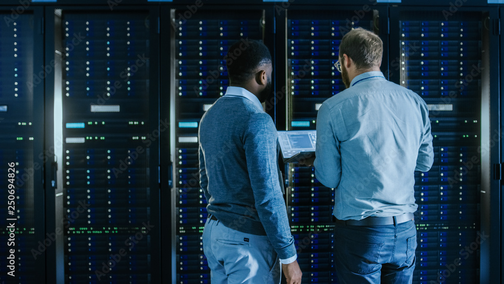 Aufkleber Bearded IT Technician in Glasses with a Laptop Computer and Black Male Engineer Colleague are Working in Data Center while Working Next to Server Racks. Running Diagnostics or Doing Maintenance Work. 