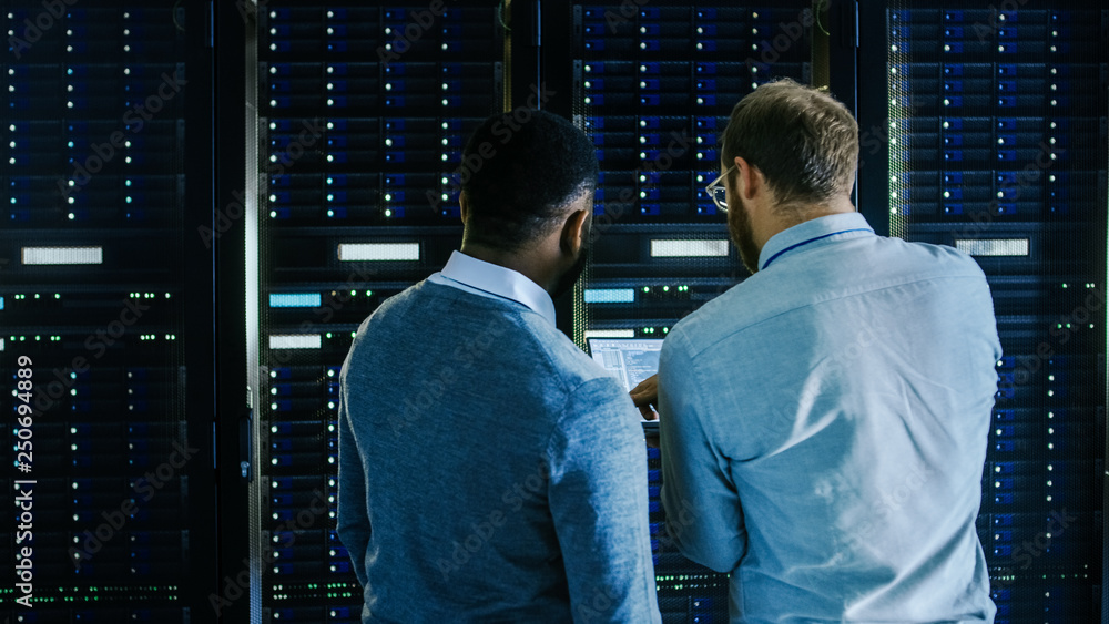 Poster Bearded IT Technician in Glasses with a Laptop Computer and Black Male Engineer Colleague are Talking in Data Center while Working Next to Server Racks. Running Diagnostics or Doing Maintenance Work. 