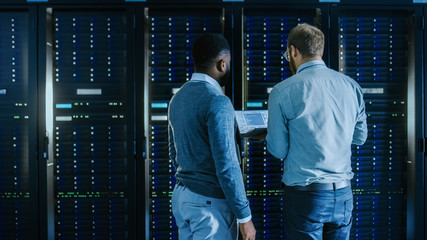 Bearded IT Technician in Glasses with a Laptop Computer and Black Male Engineer Colleague are Working in Data Center while Working Next to Server Racks. Running Diagnostics or Doing Maintenance Work. 