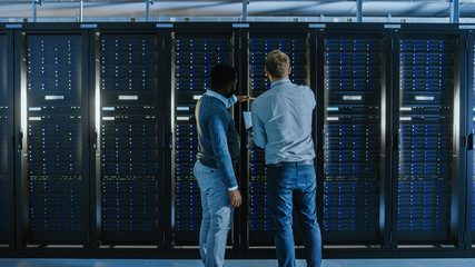 Bearded IT Technician in Glasses with a Laptop Computer and Black Male Engineer Colleague are Talking in Data Center while Working Next to Server Racks. Running Diagnostics or Doing Maintenance Work. 