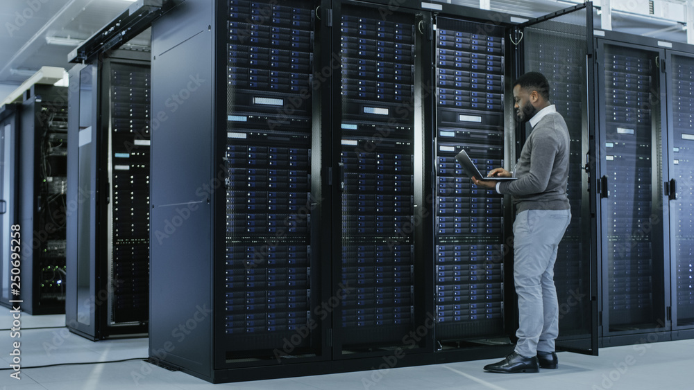 Aufkleber Black IT Specialist is Working on Laptop in Data Center while Standing Before Server Rack. Running Diagnostics, Putting in Data or Doing Maintenance Work.