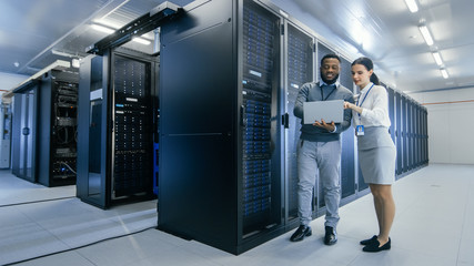 Black IT Technician with a Laptop Computer Gives a Tour to a Young Intern. They Talk in Data Center...