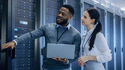 Black IT Technician with a Laptop Computer Gives a Tour to a Young Intern. They Talk in Data Center...