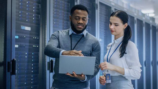 Black IT Technician with a Laptop Computer Gives a Tour to a Young Intern. They Talk in Data Center while Walking Next to Server Racks. Running Diagnostics or Doing Maintenance Work. 