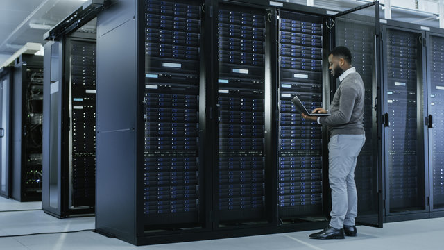 Black IT Specialist is Working on Laptop in Data Center while Standing Before Server Rack. Running Diagnostics, Putting in Data or Doing Maintenance Work.