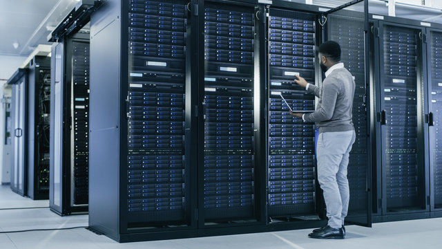 Black IT Specialist is Working on Laptop in Data Center while Standing Before Server Rack. Running Diagnostics, Putting in Data or Doing Maintenance Work.