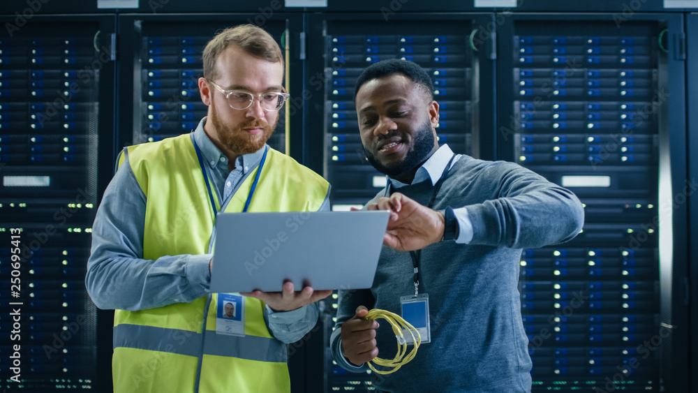 Poster Bearded IT Specialist in Glasses and High Visibility Vest with a Laptop Computer and Black Technician Colleague Talking in Data Center while Standing Next to Server Racks. Running Diagnostics. 