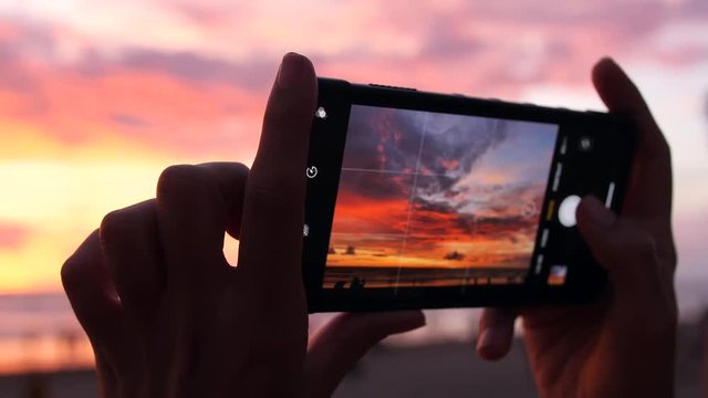 Female Hands Holding Smartphone and Doing Nature Photography. Young Tourist Woman Taking Photos with Mobile Phone Camera of Amazing Sunset at the Beach. 4K Slowmotion. Bali, Indonesia.
