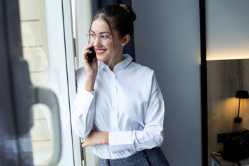Stylish young businesswoman talking with her smartphone and looking sideways standing near the window at hotel room.