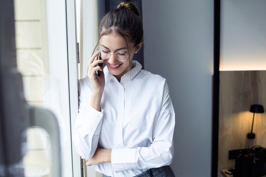 Pretty young businesswoman talking with her smartphone standing near the window at hotel room.