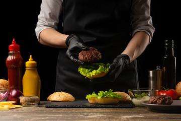 Chef puts beef patty on a burger loaf, against the background of the ingredients. Delicious and harmful food, fast food, homemade recipes, restaurant, catering, recipe book
