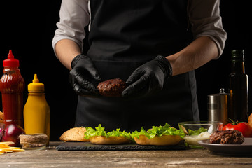 Chef puts beef patty on a burger loaf, against the background of the ingredients. Delicious and harmful food, fast food, homemade recipes, restaurant, catering, recipe book