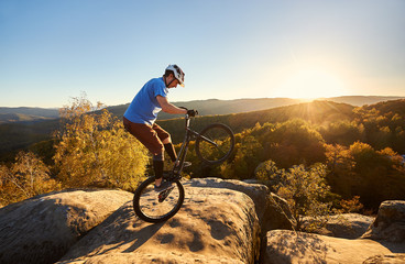 Professional cyclist riding on back wheel on trial bicycle. Sportsman rider balancing on the edge of big boulder on the top of mountain at sunset. Concept of extreme sport active lifestyle