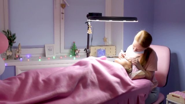 Eyelash Extension Procedure. Woman Eye with Long false Eyelashes. Girl cient lies in beauty salon. Eyelash wizard beautician with tweezers.