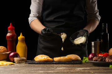 Chef puts sauce on a burger loaf, against the background of ingredients. Delicious and harmful food, fast food, homemade recipes, restaurant, catering, recipe book
