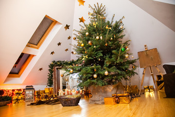 Christmas decorations in the studio, big natural fir tree with golden balls, bows and snowflakes, wooden fireplace and candles on it and brown cozy armchair