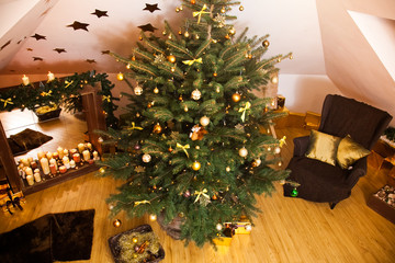 Christmas decorations in the studio, big natural fir tree with golden balls, bows and snowflakes, wooden fireplace and candles on it and brown cozy armchair