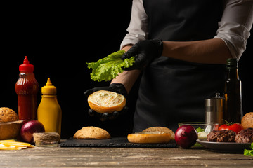 The chef puts fresh salad on a burger loaf, with the background ingredients. Delicious and harmful food, fast food, homemade recipes, restaurant, catering, recipe book