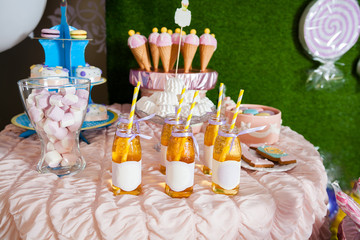Candy bar for the birthday for little baby girl. Sweet table and nice ice-creams from sugar horn and pink and white icing, bottles with juice, small colored macaroons, different cakes. Green backgroun