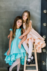 two beautiful little girls posing on a grey background , one of them is sitting on a wooden chair, and the other hugs her