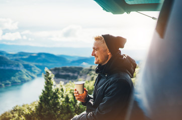 hipster tourist hold in hands mug of hot drink, lonely guy smile enjoy sun flare mountain in auto,...