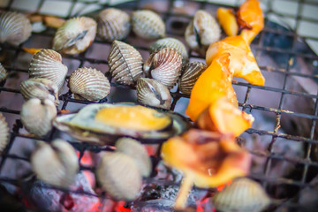 Grilled squids, mussels and clams on charcoal stove at night party. Seafood barbecue party with squid calamaris, mussels and clams are grilling on fire. BBQ concept, selective focus.