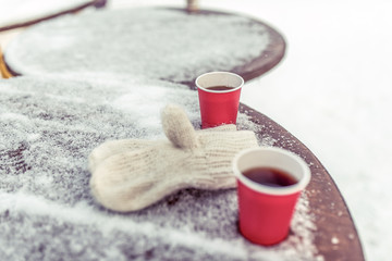 Fototapeta na wymiar Close-up wooden table in cafe covered with snow. On table there are warm white mittens, two red cups of coffee and tea. The concept of snack and dinner winter holiday resort. Free space for text.